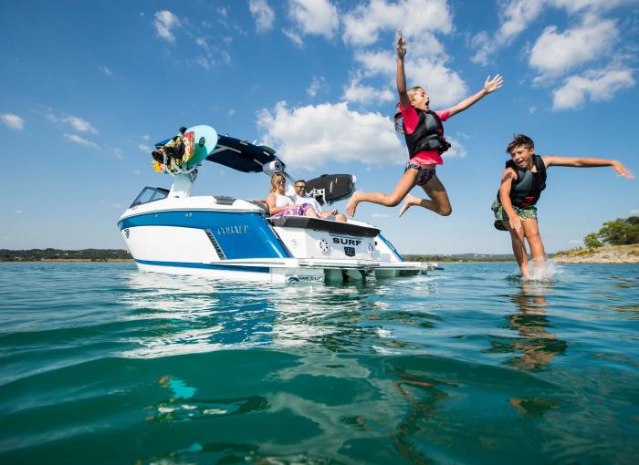 Kids jumping off of a blue and white Cobalt Surf Boat while parents sit back and watch